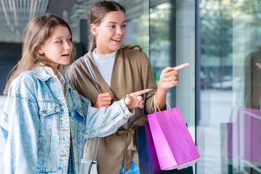Two caucasian teenage girls enjoy shopping, smiling and pointing at a store window while holding a bright pink bag, reflecting their excitement and interest during a casual outing.