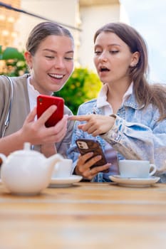 Two teenage girls share a fun moment at an outdoor cafe, smiling and pointing at smartphones. Surrounded by tea cups and sunshine, they enjoy friendship and technology together. Vertical