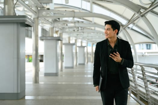 Adult businessman using mobile phone walking outdoors in a modern city walkway.