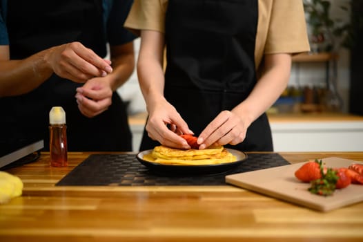 Close up of couple decorating pancakes with fresh strawberries in a kitchen.
