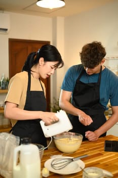 Young couple in aprons preparing pancake batter and following a recipe on digital tablet.