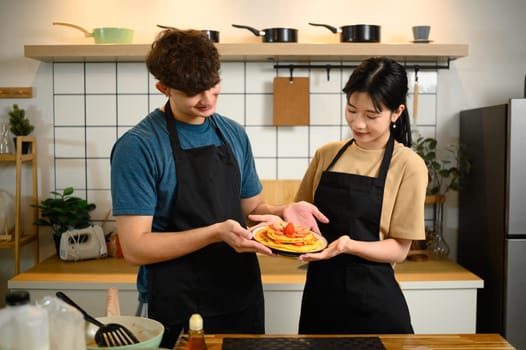 Young couple proudly presenting a plate of homemade pancakes topped with strawberries.