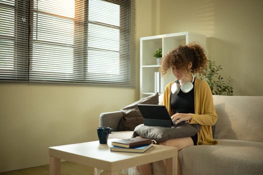 Young woman sitting on a sofa with headphones around her neck and using digital tablet.