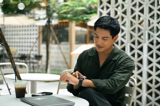 Asian man checking smartwatch while sitting at outdoor cafe table, with iced coffee and laptop nearby.