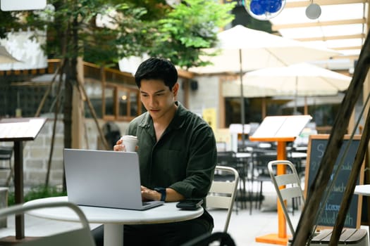 Smiling man drinking coffee and using on a laptop at a modern outdoor cafe.