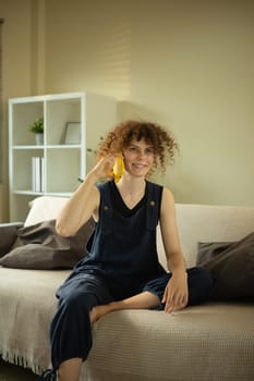 Happy young woman with curly hair talking on the phone sitting in bright living room.