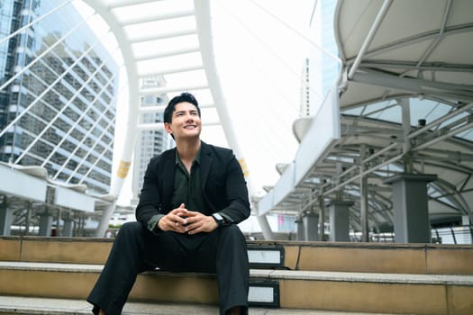 Confident businessman sitting on steps in urban cityscape and looking up with optimism.