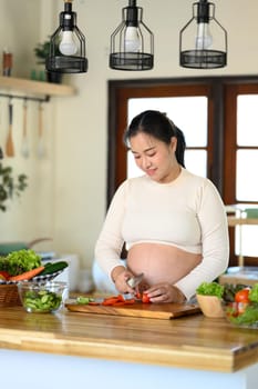Happy pregnant woman preparing a nutritious meal with fresh ingredients in home kitchen.