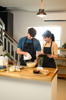 Young couple in aprons preparing pancake batter and following a recipe on digital tablet.