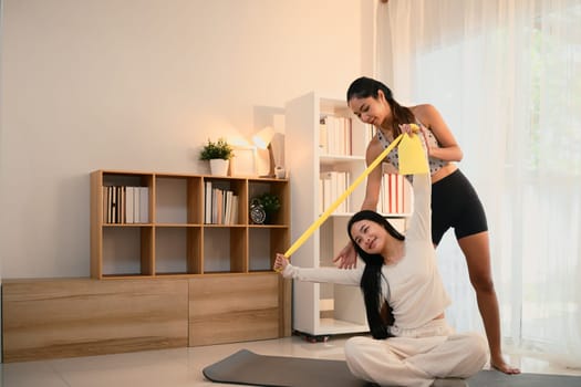 Active mother helping her daughter stretch with a resistance band in a bright living room.