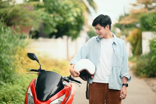 Young asian man holding a helmet standing next to his scooter in a suburban neighborhood.