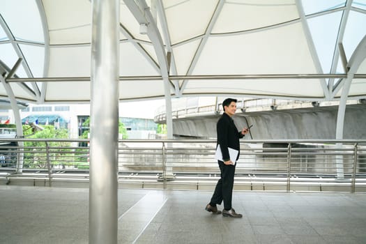 Asian businessman walking through modern transit walkway, holding smartphone and laptop.