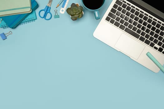 Top view of modern workspace with laptop, stationery and coffee cup on pastel blue background.