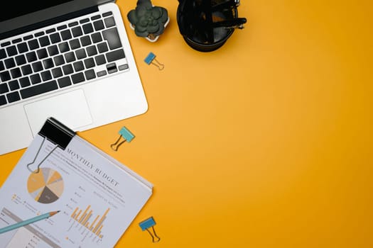 Modern office desk with laptop, monthly budget report, stationery, and succulent on yellow background.