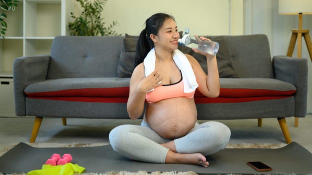 Happy pregnant woman relaxing after exercise, sitting on a yoga mat and drinking water.