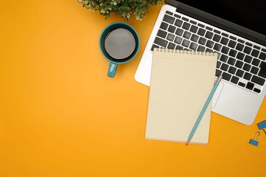 Flat lay of modern desk with a coffee cup, laptop and notebook on yellow background.