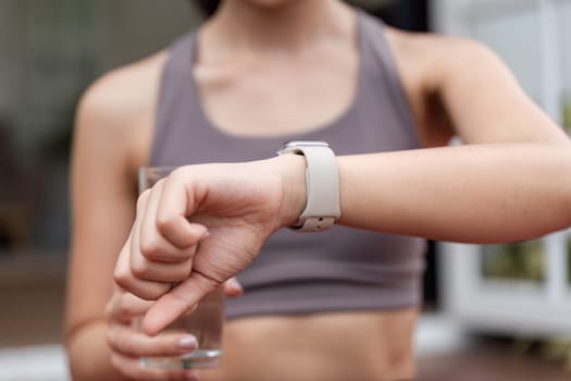 A young woman checks her smartwatch for fitness updates, holding a glass of water, emphasizing health and hydration.