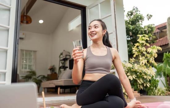 A woman smiling and drinking water in a yoga pose, highlighting hydration and wellness in her home environment.