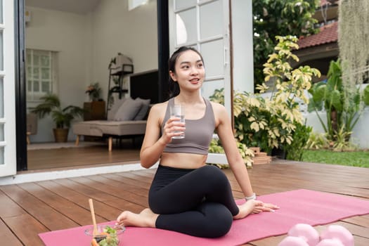 A woman sitting in a yoga pose, drinking water and promoting hydration as part of her wellness routine in a serene home environment.