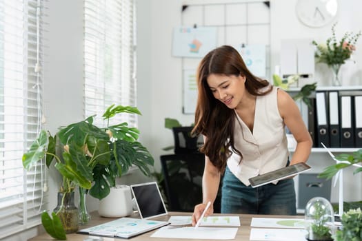 A focused young woman engaged in business sustainability planning, surrounded by greenery in an eco-friendly office environment.