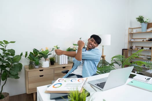 A young man stretches and smiles in his home office, surrounded by lush indoor plants, showcasing the benefits of incorporating greenery into work-life balance for sustainability.