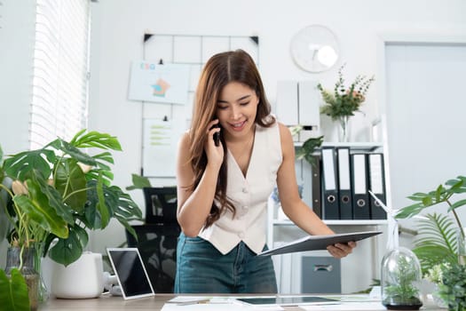 A young businesswoman smiling as she reviews reports and speaks on the phone in a modern office setting, fostering communication and teamwork.