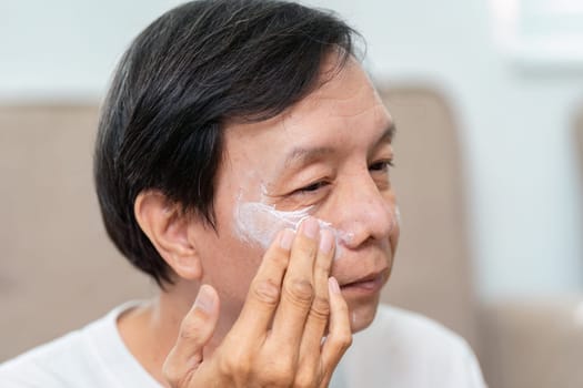 An elderly man gently applies moisturizer to his face, emphasizing self-care and skincare routines for mature skin in a comfortable home environment.