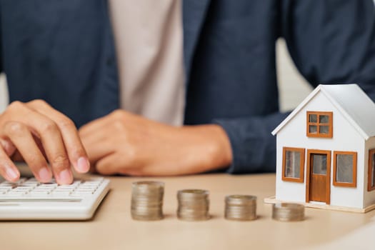 Person using calculator next to house model and cash stacks. Reflects real estate and property tax planning, profit increase strategies, and financial decisions for people aiming for smart investments