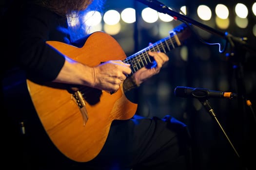 Close-up of a musician playing an acoustic guitar on a stage with microphones and lights in the background