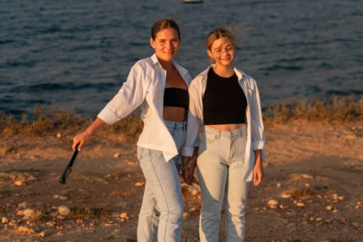 Two young women wearing white shirts and jeans stand on a beach. One holds a black object in her hand. The ocean is behind them.