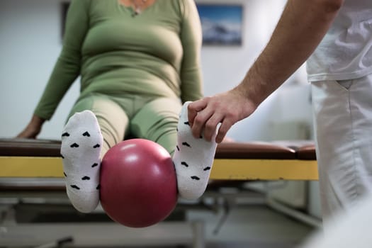 Close up of a patient performing a physical therapy exercise using a ball between the legs, assisted by a therapist.
