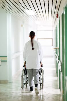 A smiling young man in a wheelchair is being pushed by a female healthcare worker in a white coat and stethoscope. They are in a hospital corridor, suggesting a medical or rehabilitation environment, evoking care and recovery.