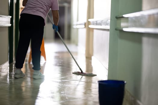 Female janitor standing in a hospital corridor with mop and bucket, symbolizing cleanliness and hygiene.