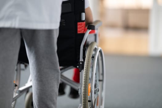 A smiling young man in a wheelchair is being pushed by a female healthcare worker in a white coat and stethoscope. They are in a hospital corridor, suggesting a medical or rehabilitation environment, evoking care and recovery.