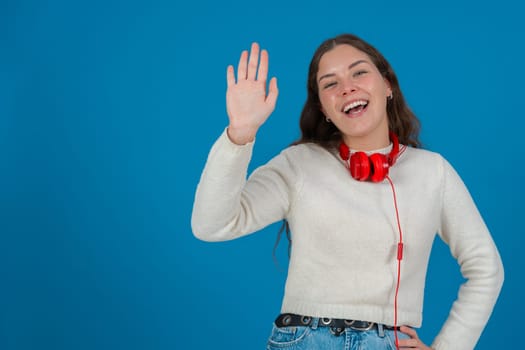 Happy young woman wearing a white sweater and red headphones, waving at the camera with a cheerful expression, standing against a vibrant blue background, symbolizing greeting and joy. Copy space