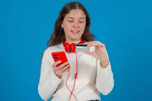 Young woman with red headphones holding a credit card and smartphone while shopping online. She looks focused and calm, standing in front of a blue background during a digital transaction.