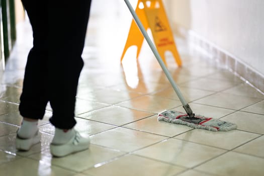 A janitor mopping a tiled floor in a brightly lit hallway with a visible caution sign, highlighting cleanliness, hygiene, and workplace maintenance.