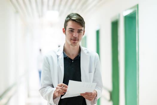 Young male doctor walking in hospital hallway while reading patient documentation. Clinical work environment.
