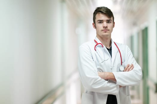 Portrait of a young doctor in white coat with arms crossed, standing in a hospital hallway.