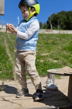 Young boy wearing a helmet enjoys dropping sand through his fingers at a playground on a sunny day