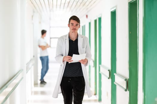 Young male doctor walking in hospital hallway while reading patient documentation. Clinical work environment.
