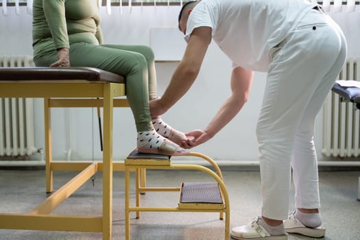 Physiotherapist helping an elderly woman with leg movement on a step platform during a rehabilitation session.
