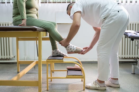 Physiotherapist helping an elderly woman with leg movement on a step platform during a rehabilitation session.