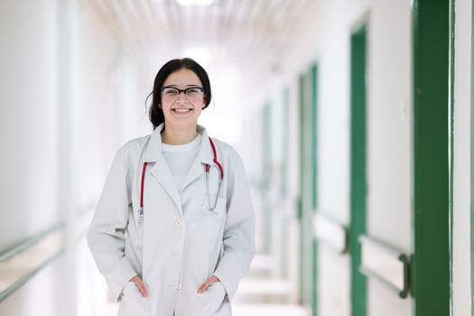 Confident young female doctor with a stethoscope smiling while standing in a bright hospital corridor.