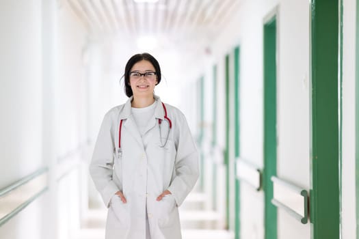Confident young female doctor with a stethoscope smiling while standing in a bright hospital corridor.