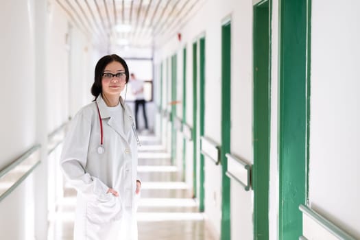 Confident young female doctor with a stethoscope smiling while standing in a bright hospital corridor.