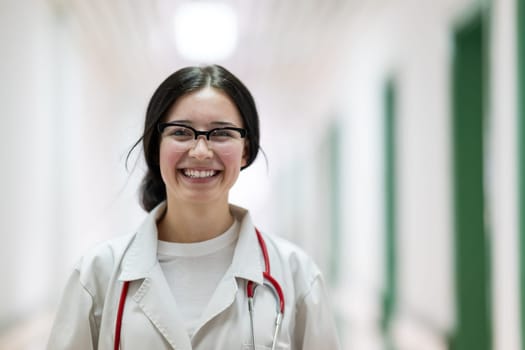 Confident young female doctor with a stethoscope smiling while standing in a bright hospital corridor.