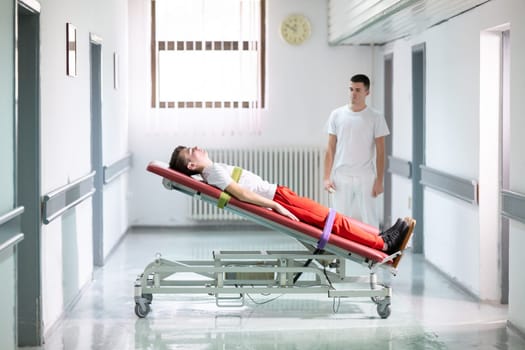 Male therapist assists patient with a standing frame during a rehabilitation session in a clinical hallway. Physical recovery support concept.