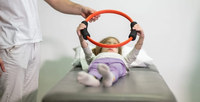 Young girl performing rehabilitation exercises with the help of a trainer, using a resistance ring while lying on a medical bench.