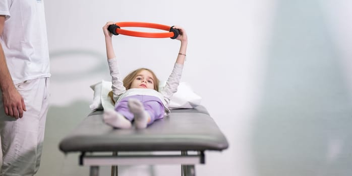 Young girl performing rehabilitation exercises with the help of a trainer, using a resistance ring while lying on a medical bench.